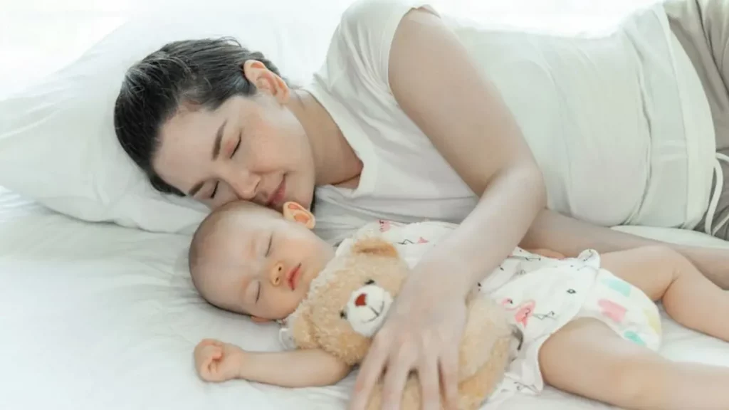 Seorang ibu tidur di samping bayi yang juga sedang tidur bersama dengan sebuah boneka teddy bear.