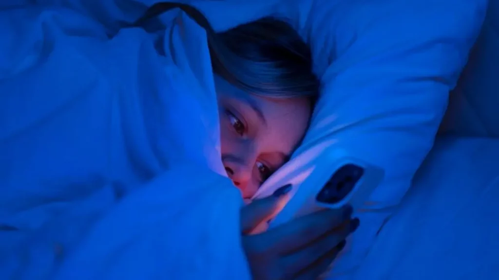 Seorang perempuan tidur menyamping dengan selimut diangkat sampai ke wajahnya sambil menggunakan ponsel.
