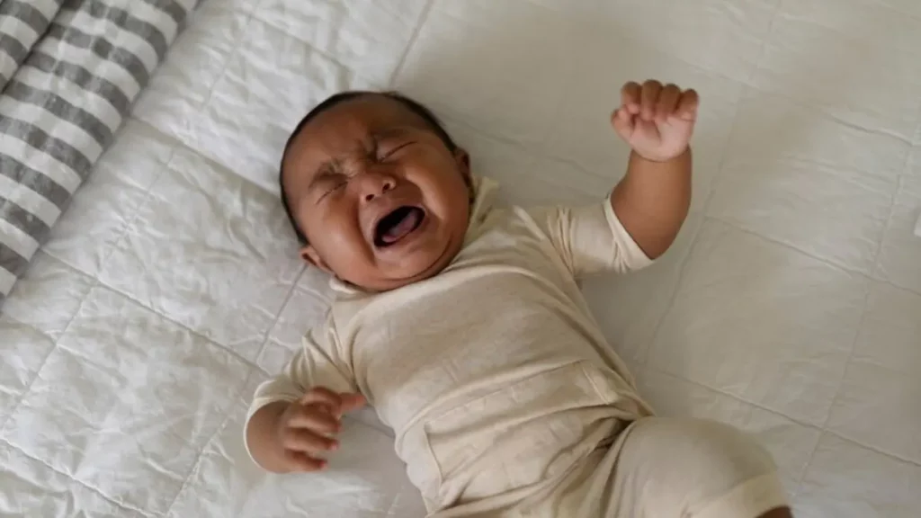 Bayi tidur telentang sambil menangis di atas kasur dengan cover warna putih.