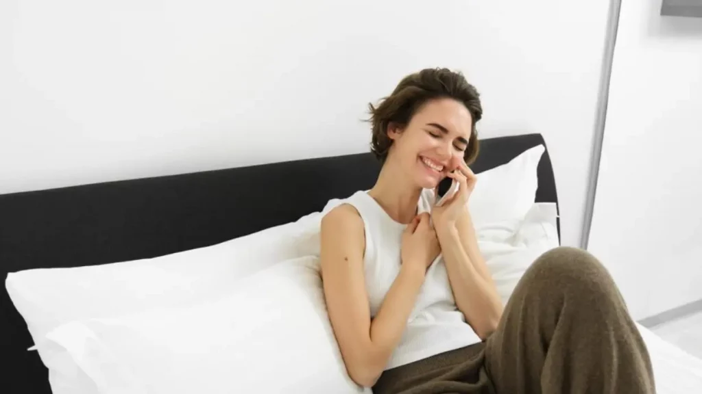 Seorang perempuan bersandar di tempat tidurnya, tersenyum, sambil melakukan panggilan telepon.