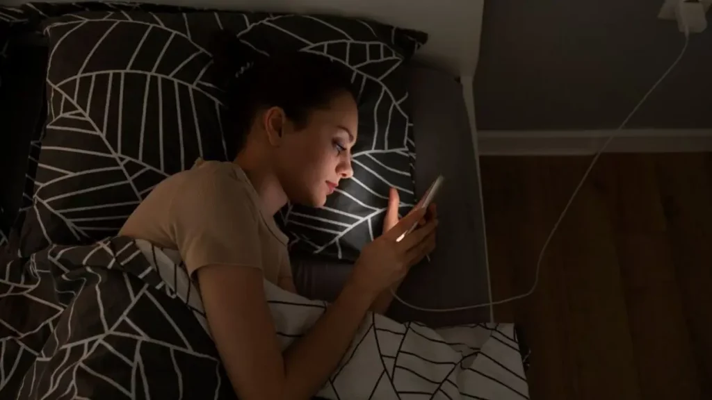 Seorang perempuan dalam posisi tidur menyamping menghadap arah kiri sambil menggunakan ponsel yang terhubung dengan pengisi daya.